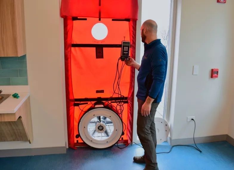 Mann bedient ein Blower-Door-Messgerät an einer roten Türabdichtung
